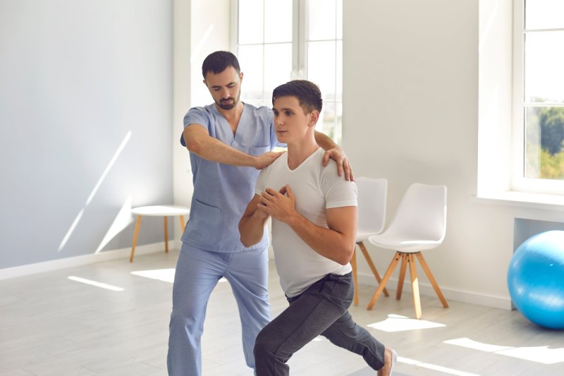 Medical center specialist helping young man recover after trauma. Professional physiotherapist helping male athlete do forward lunges exercise during physiotherapy rehabilitation after sports injury