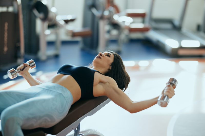 An attractive bodybuilder woman is lifting dumbbells while lying down on the bench in the gym.