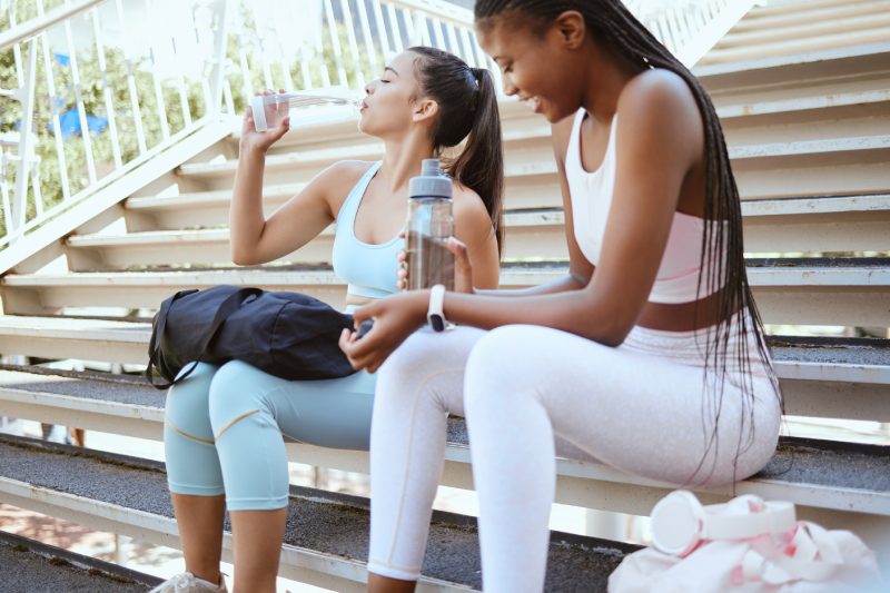 Water, fitness and friends training on stairs in the city of Sweden for workout in summer. African athlete and black woman runner drinking from bottle for energy after exercise together