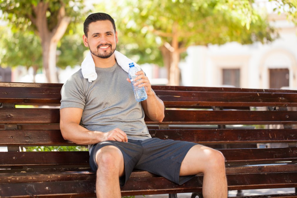 Portrait of a happy young man drinking some water from a bottle while sitting and resting in a park bench after doing some jogging outdoors